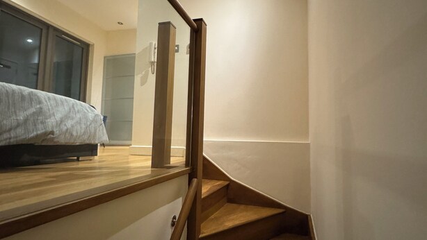 Una escalera moderna con peldaños de madera y barandilla de vidrio, que lleva a un dormitorio con cama y ventana.