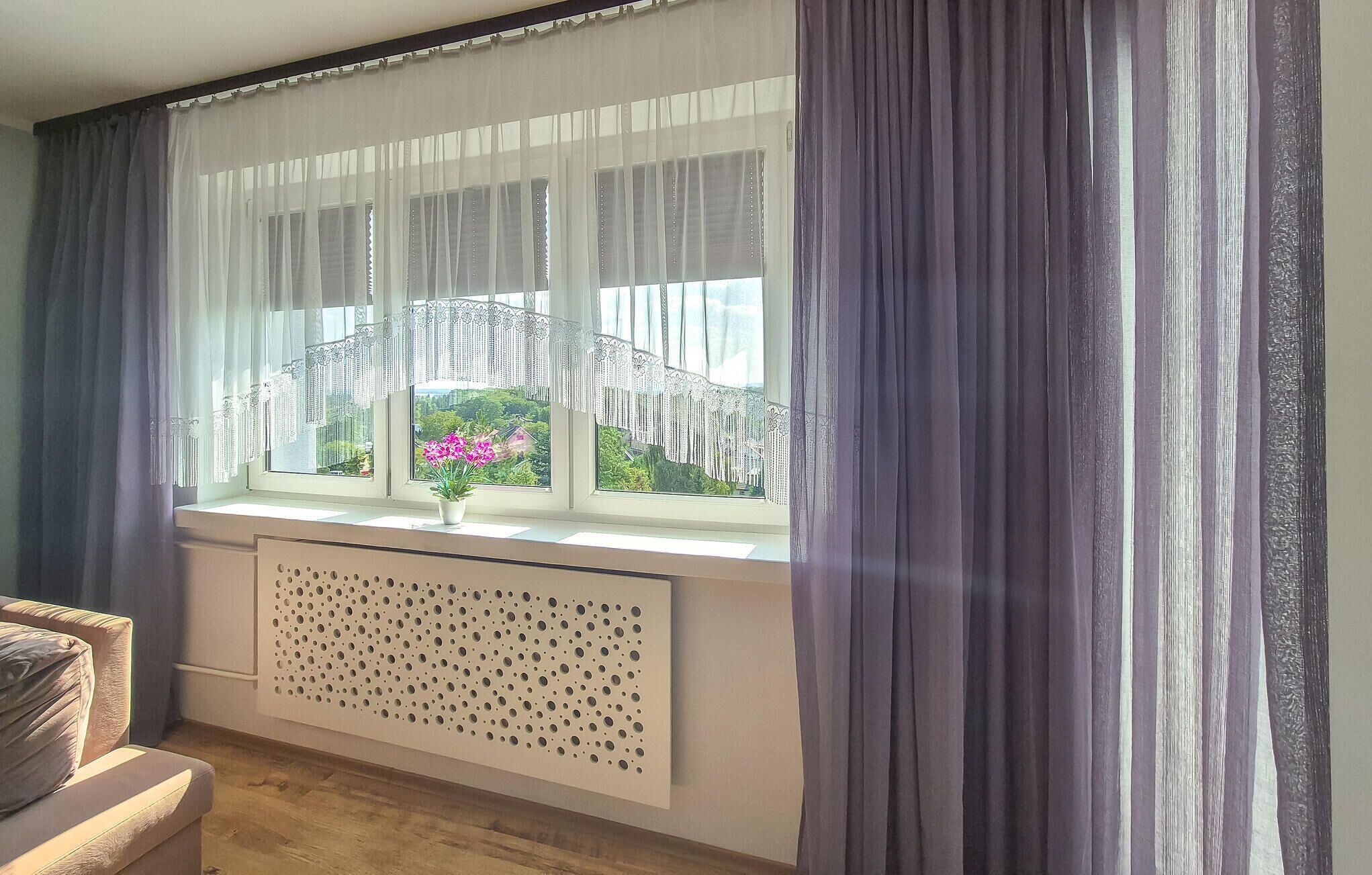 Une pièce dotée d’une grande fenêtre, de rideaux blancs et d’un radiateur.