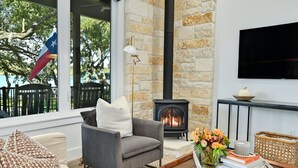 A cozy living room with a stone fireplace, a comfortable sofa, and a large window overlooking a deck with a flag. - Packsaddle Point Luxury Lakefront w/Pool+Guest House (Kingsland)