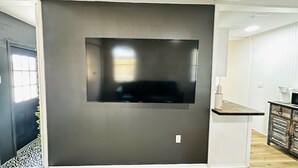 A modern living room with a large flat-screen TV mounted on a dark wall, a wooden floor, and a kitchen area with a white countertop and cabinets.