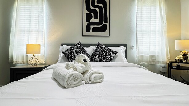 A neatly made bed with rolled towels, black and white patterned pillows, and a large abstract artwork on the wall.