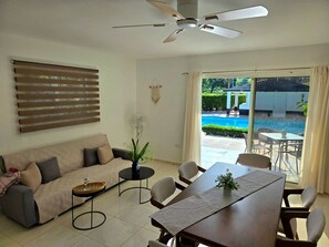 A living room with a sofa, coffee table, dining table, chairs, a ceiling fan, and a view of a swimming pool through large glass doors.