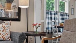 Een woonkamer met een grijze bank, een houten salontafel en een lamp. Er is een eethoek met een tafel en stoelen, en een raam met gordijnen.