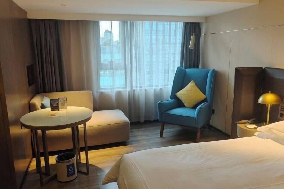 Une chambre d’hôtel avec un lit, un fauteuil bleu, une petite table, une lampe et une vue sur la ville par la fenêtre.