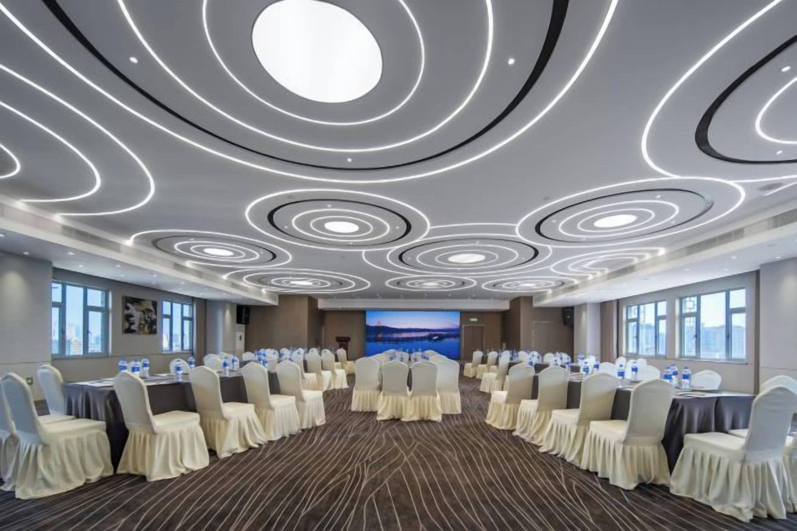 Un salón de conferencias con distribución circular, mesas y sillas redondas, y una pantalla grande al frente.