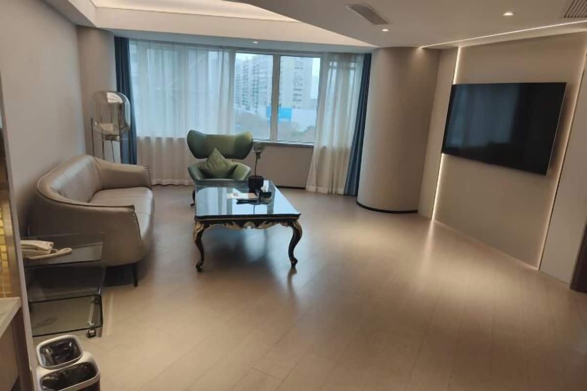 Un salon moderne avec un canapé, une table basse en verre, un téléviseur fixé au mur et une fenêtre avec des rideaux.