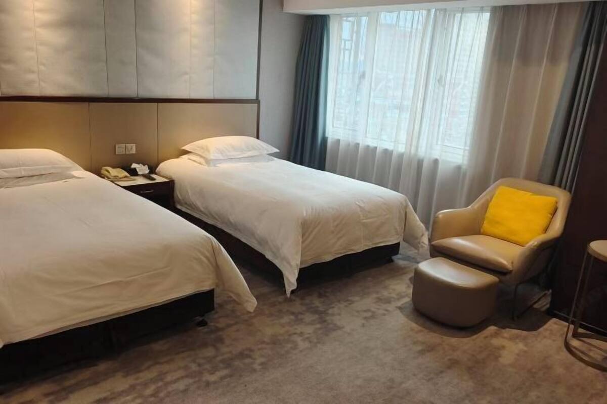 Une chambre d’hôtel avec deux lits, une chaise avec un coussin jaune, une table de chevet avec un téléphone et une grande fenêtre avec des rideaux.