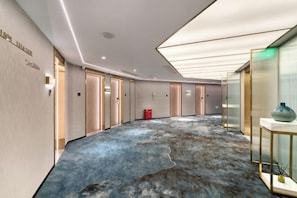 Un pasillo de hotel con una alfombra azul distintiva, puertas y un jarrón sobre una mesa.