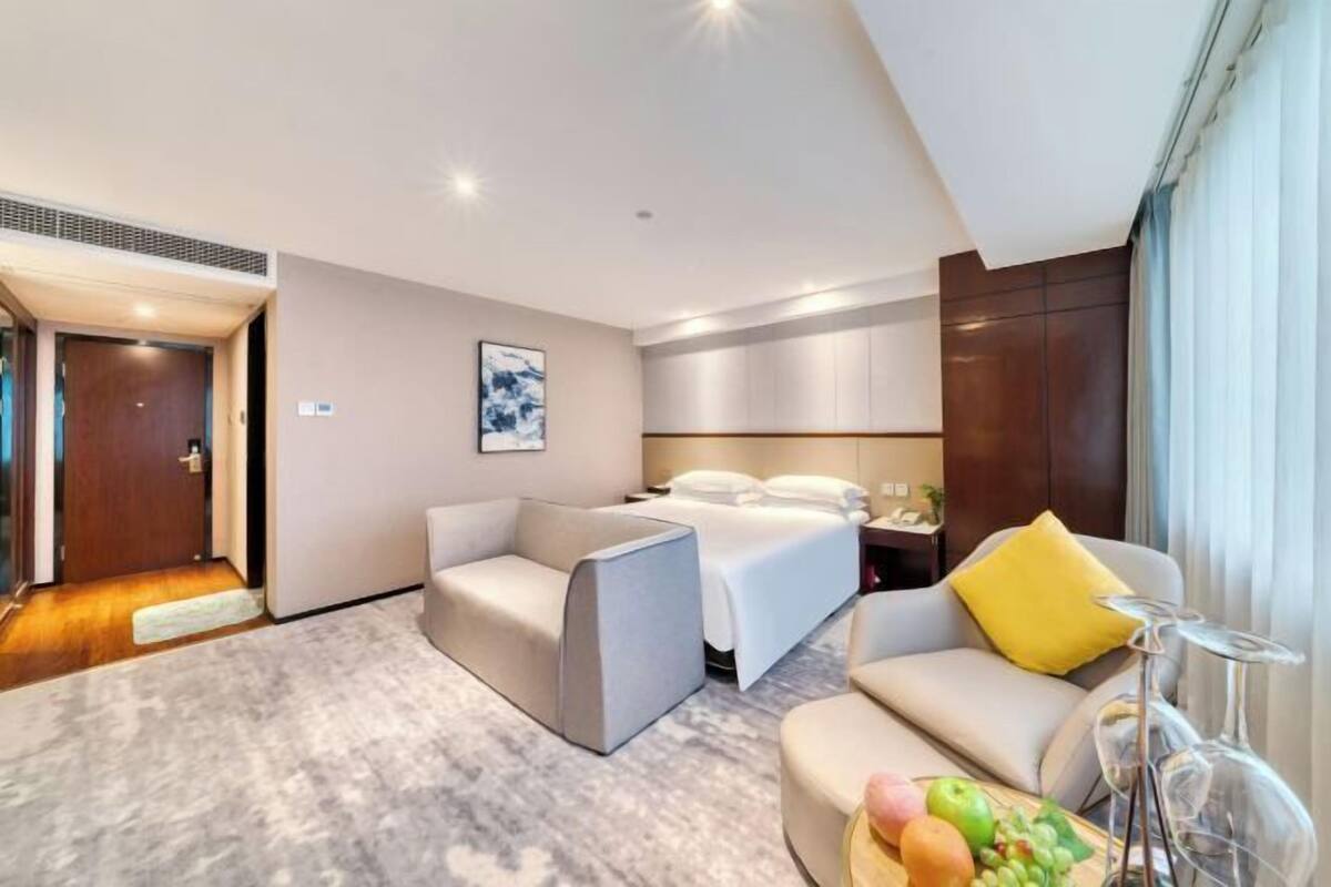Une chambre d’hôtel moderne avec un lit, deux canapés, une table basse avec des fruits et une grande fenêtre avec des rideaux.