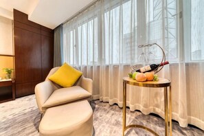 Ein modernes Wohnzimmer mit einer Couch, einem kleinen runden Tisch mit einer Flasche und Obst sowie einem großen Fenster mit Vorhängen.