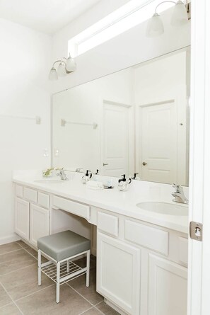 Un baño con tocador blanco, dos lavamanos, un espejo grande y un taburete.