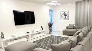 Ein modernes Wohnzimmer mit Flachbildfernseher, grauem Sofa, Hocker, weißem Unterhaltungsschrank und einem gemusterten Teppich.