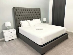 Un dormitorio moderno con cabecera capitonada, una cama grande con ropa blanca, una mesita de noche con lámpara y una cortina oscura.
