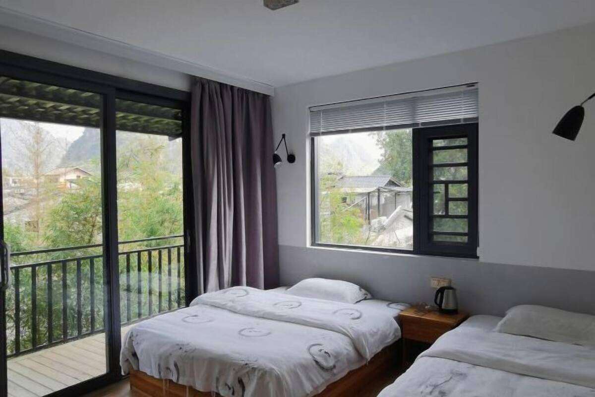 Une chambre avec une grande fenêtre, un balcon, un lit, une table de chevet et une lampe.