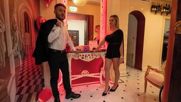 Un homme et une femme se tiennent dans une pièce dont le sol est recouvert d’un motif à carreaux rouges et blancs, où se trouve une table richement décorée, également aux couleurs rouge et blanc, et un lustre.