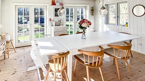 A white dining table with wooden chairs, a vase of flowers, and a view of the outdoors through large windows. - Pet friendly home in Sjællands Odde (Sjællands Odde)