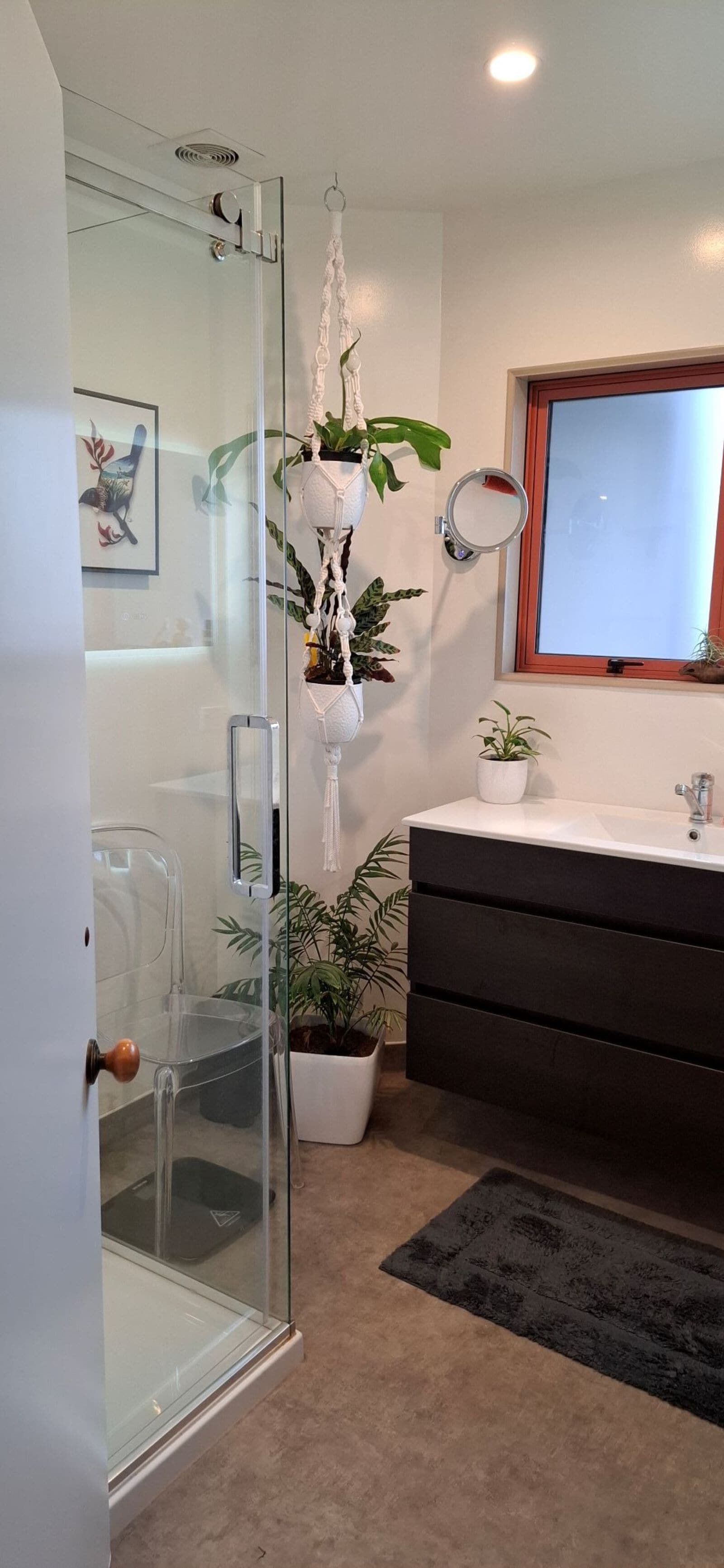 A modern bathroom with a glass shower enclosure, a white vanity with a dark drawer unit, a potted plant, and a framed artwork on the wall.