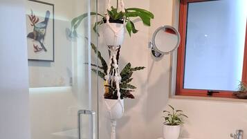 A modern bathroom with a glass shower enclosure, a white vanity with a dark drawer unit, a potted plant, and a framed artwork on the wall.