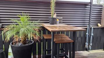 A small outdoor seating area with a wooden table and chairs, surrounded by potted plants and a metal fence.