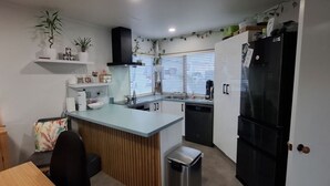 A compact kitchen with a central island, a dishwasher, a refrigerator, and a dining area with a table and chairs. - Townhouse Haven - Modern, Spa, close to Nelson Central! Pets welcome. (Nelson)