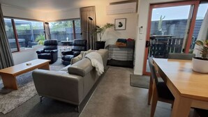 A modern living room with a sofa, two armchairs, a coffee table, and a dining table. There is a large window providing a view of the backyard. - Townhouse Haven - Modern, Spa, close to Nelson Central! Pets welcome. (Nelson)