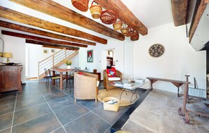 A spacious living area with a high ceiling, exposed wooden beams, a dining table with chairs, a wicker sofa, and a small table with books and a basket.