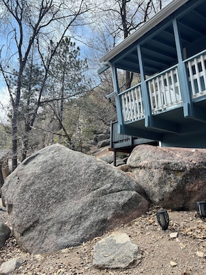 Eine Holzhütte mit blauer Fassade und weißen Geländern, umgeben von großen Felsen und Bäumen.
