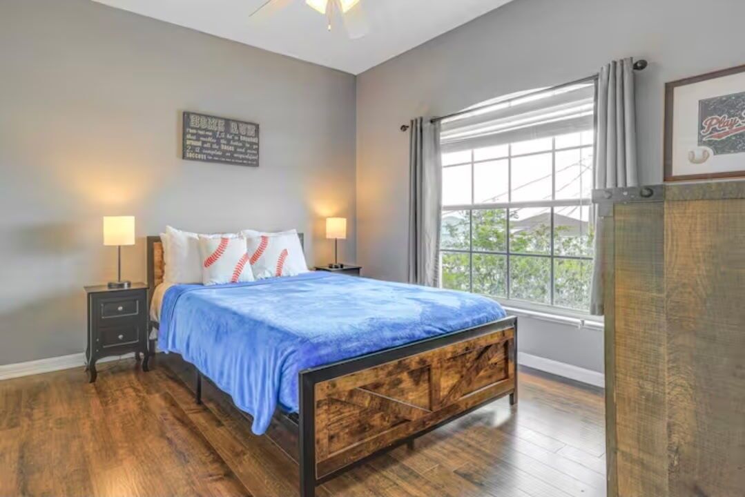 A bedroom with a wooden bed, two bedside tables, a window with curtains, and a wall-mounted artwork.