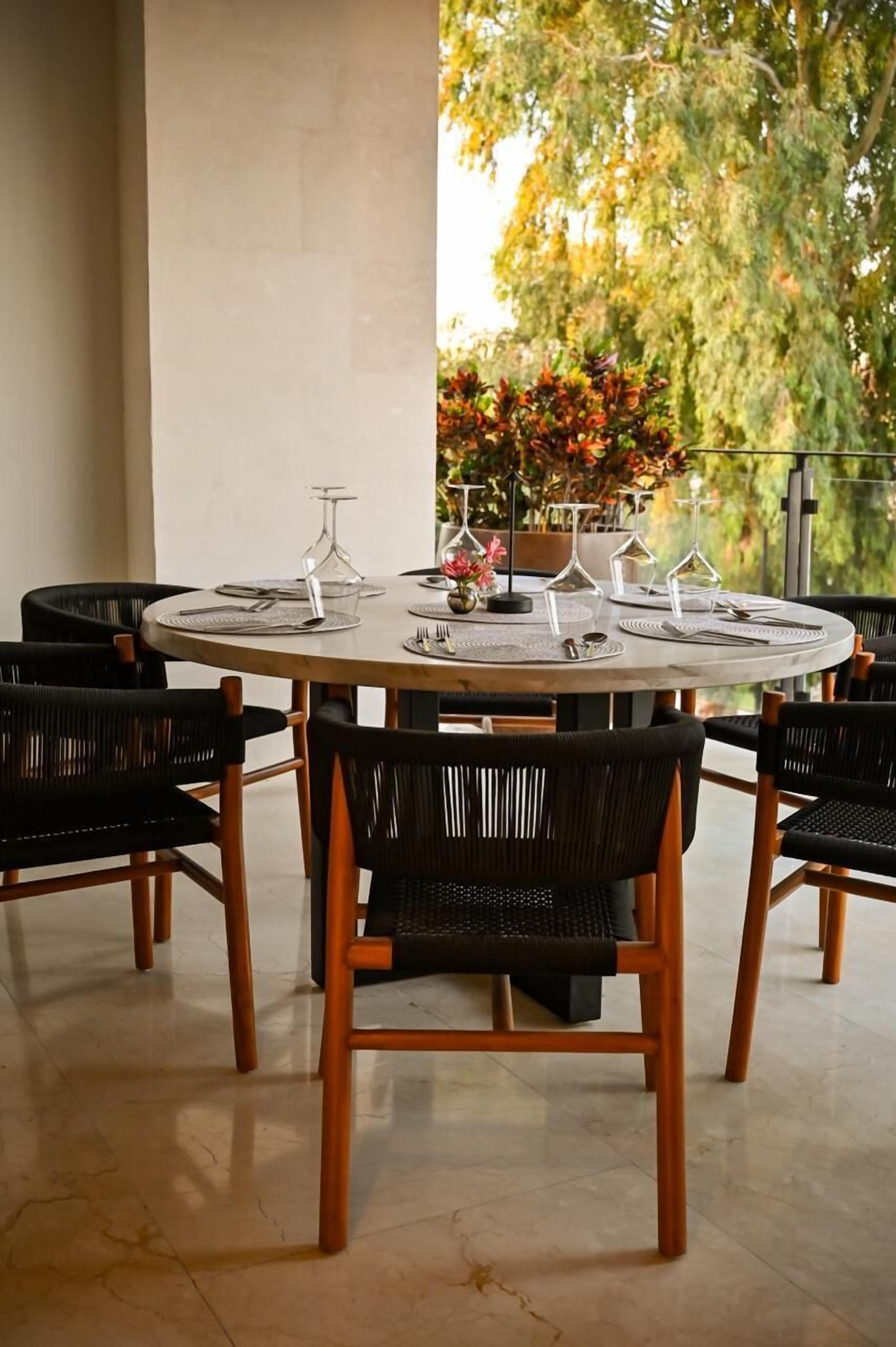 A round dining table with six chairs, set with place settings and wine glasses, surrounded by a balcony with a view of trees.