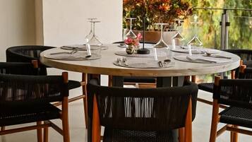 A round dining table with six chairs, set with place settings and wine glasses, surrounded by a balcony with a view of trees.