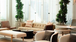 Una sala moderna con un sofá beige, una mesa de centro y una mesa de comedor. Hay dos plantas en macetas y una ventana grande con persianas.
