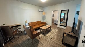 A modern living room with a brown leather sofa, a wooden coffee table, a desk, and a chair. There is a TV on a wooden stand and a mirror on the wall. - Central ICT.B (Wichita)