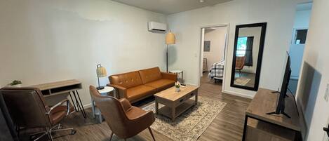 Una sala de estar moderna con un sofá de cuero marrón, una mesa de centro de madera, un escritorio y una silla. Hay un televisor sobre un mueble de madera y un espejo en la pared.