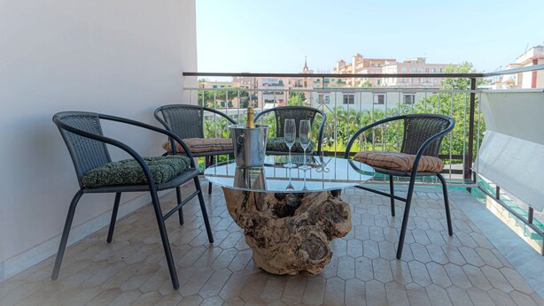 Ein Balkon mit einem runden Glastisch, vier Korbstühlen und einer Metallvase mit Gläsern.