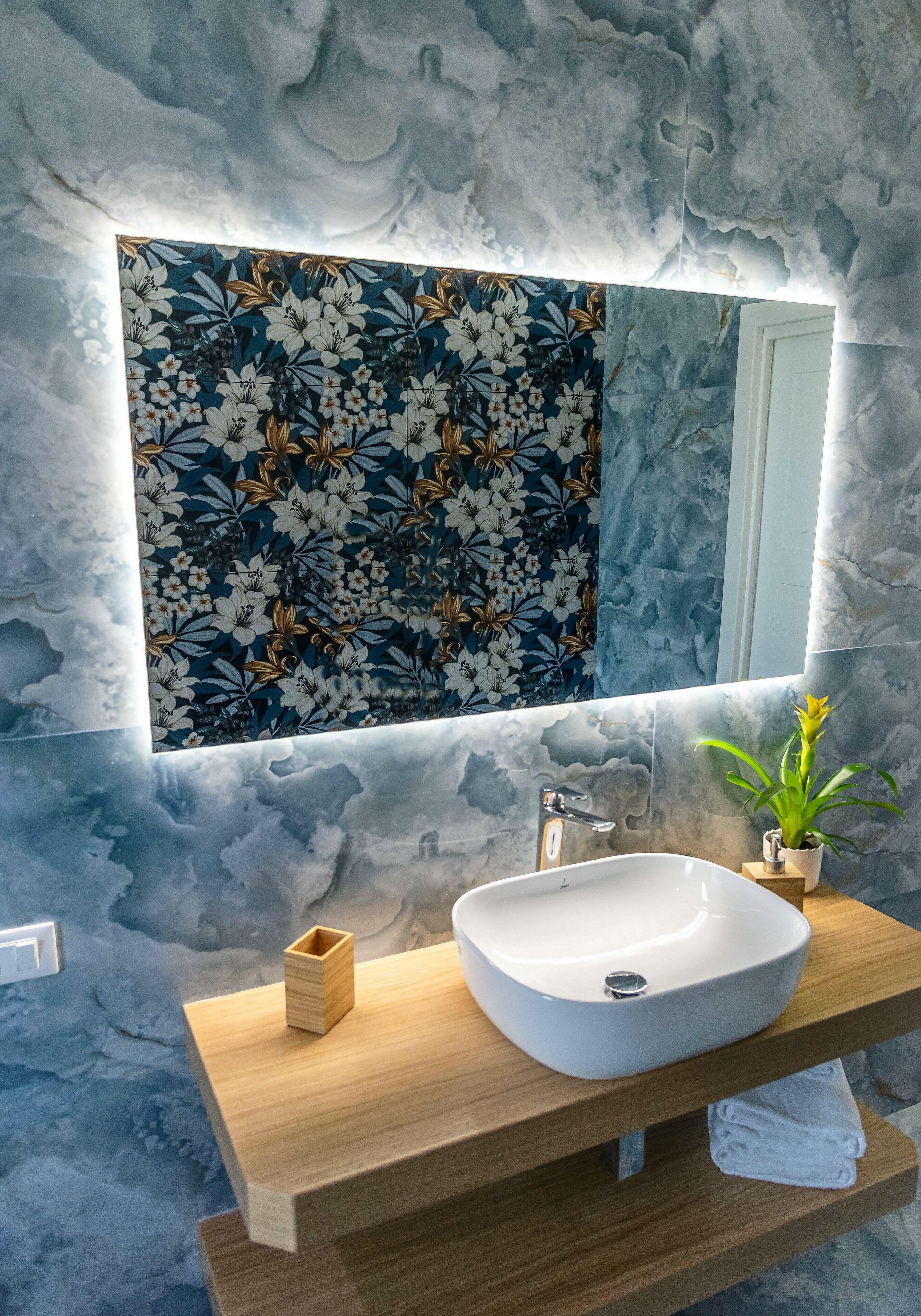 A bathroom with a marble wall, a floral-patterned mirror, a white sink, and a small potted plant.