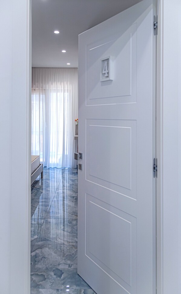 Un pasillo con una puerta blanca numerada 4, que conduce a una habitación con cama y ventana con cortinas traslúcidas.