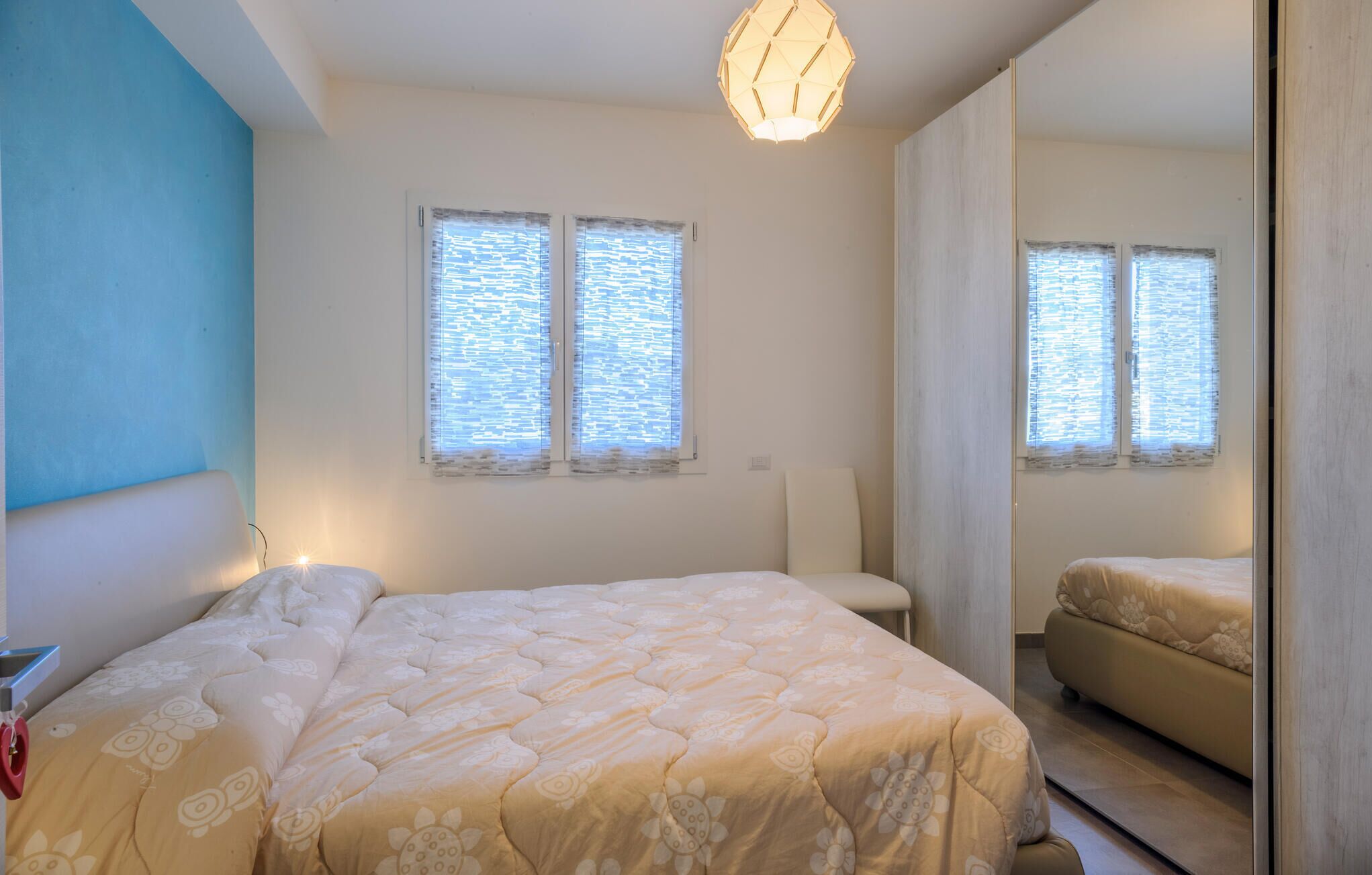 Un dormitorio con una cama, una silla y un espejo. Hay dos ventanas con persianas.