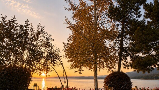 An outdoor seating area with a fire pit, chairs, and tables, overlooking a body of water during sunset.