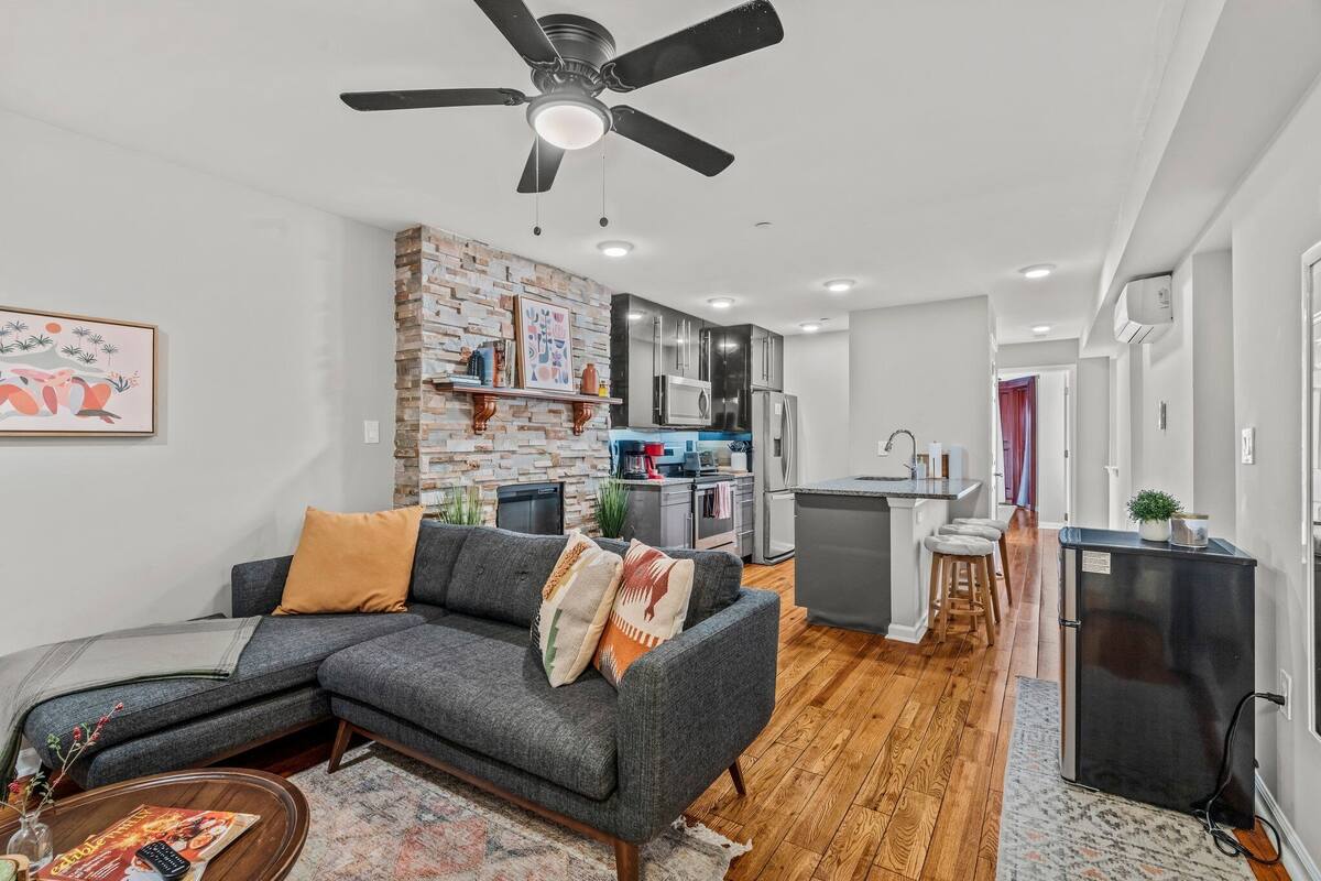 A modern living room with a grey sectional sofa, a ceiling fan, and a kitchen in the background.