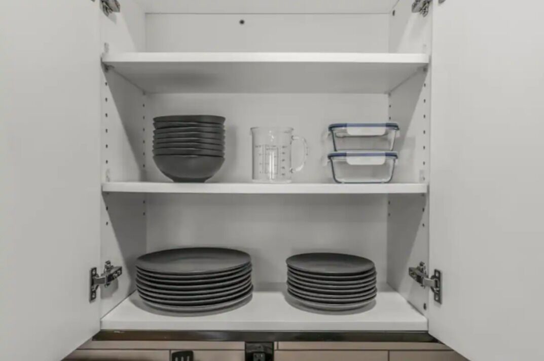 A kitchen cabinet with two shelves, each holding stacks of plates and bowls, and a measuring cup.