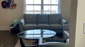 A living room with a gray sofa, a glass coffee table, and a window with a view of trees. - Pleasant 4-bedroom house in New Roads with WiFi. (New Roads)
