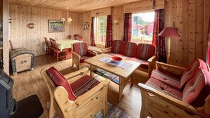 A wooden cabin interior with a dining table, chairs, a sofa, and a small chest of drawers.