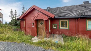 A red wooden house with a sloped roof, a wooden door, and two windows with green frames.