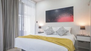 A hotel room with a bed, two bedside tables, a nightstand, a large abstract painting, and a window with curtains.