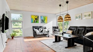Una sala moderna con un sofá de cuero negro, un sofá gris y un sillón negro. Hay dos cuadros en la pared, un televisor montado y un amplio ventanal con vistas a la vegetación.
