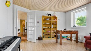 Una habitación amplia con piso de madera, un piano, una estantería, una mesa de fútbol de mesa y una ventana que ofrece vistas a árboles.