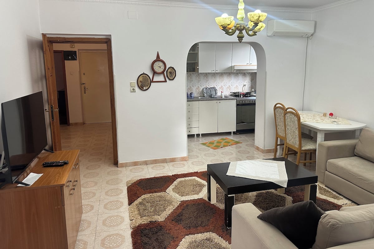 A living room with a TV, a small table, a sofa, and a kitchen in the background.