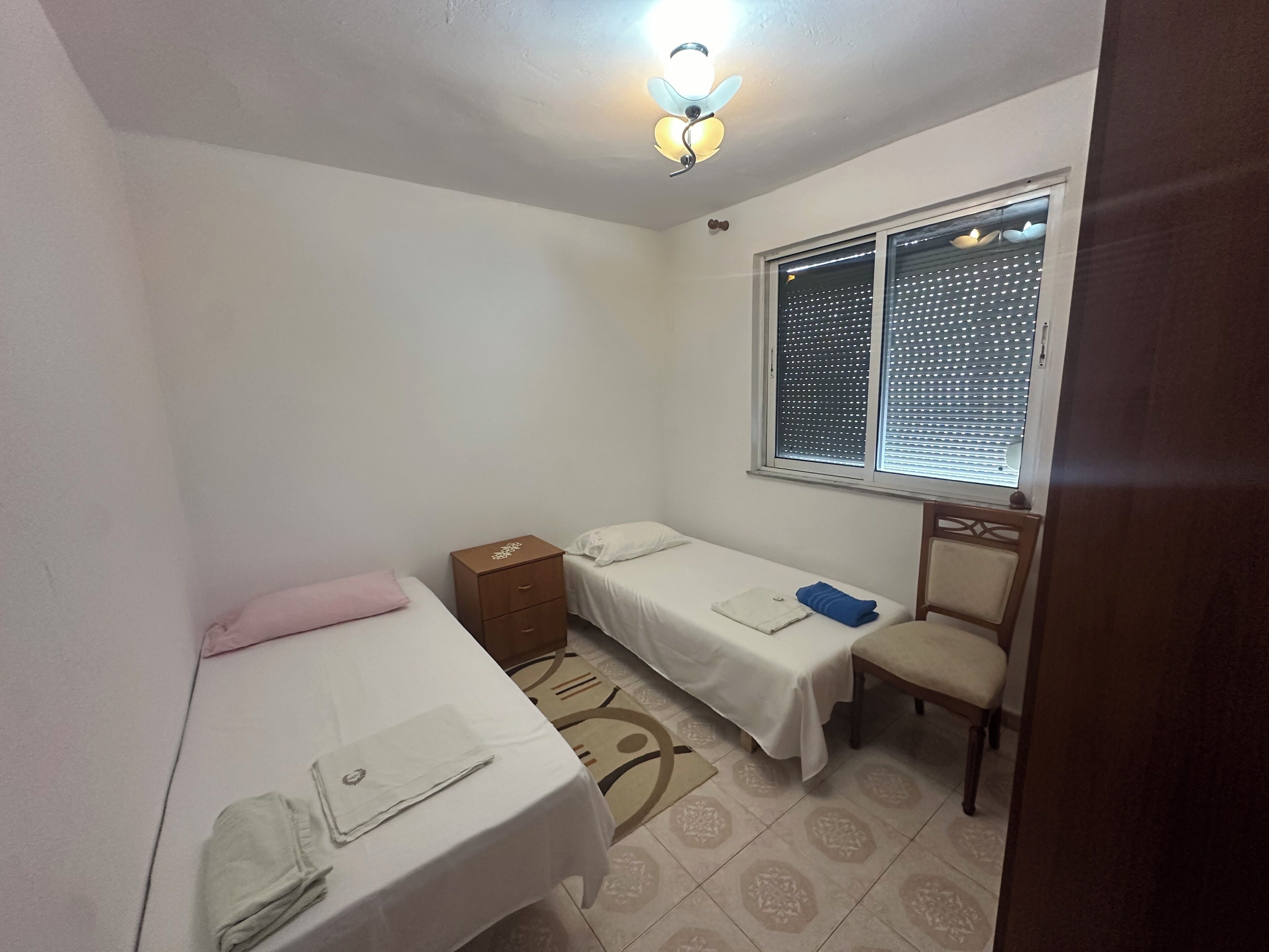 A small, clean room with two single beds, a wooden nightstand, a chair, and a window with blinds.