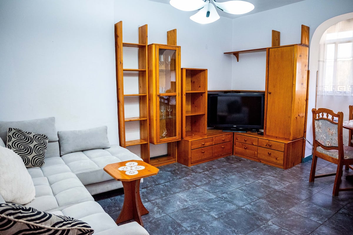 A living room with a sofa, a coffee table, wooden shelving, a television, and a dining area with a chair.