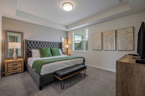 Un dormitorio con cabecera capitonada, cama con ropa de cama verde, mesita de noche con lámpara, una banca y un escritorio con una computadora.